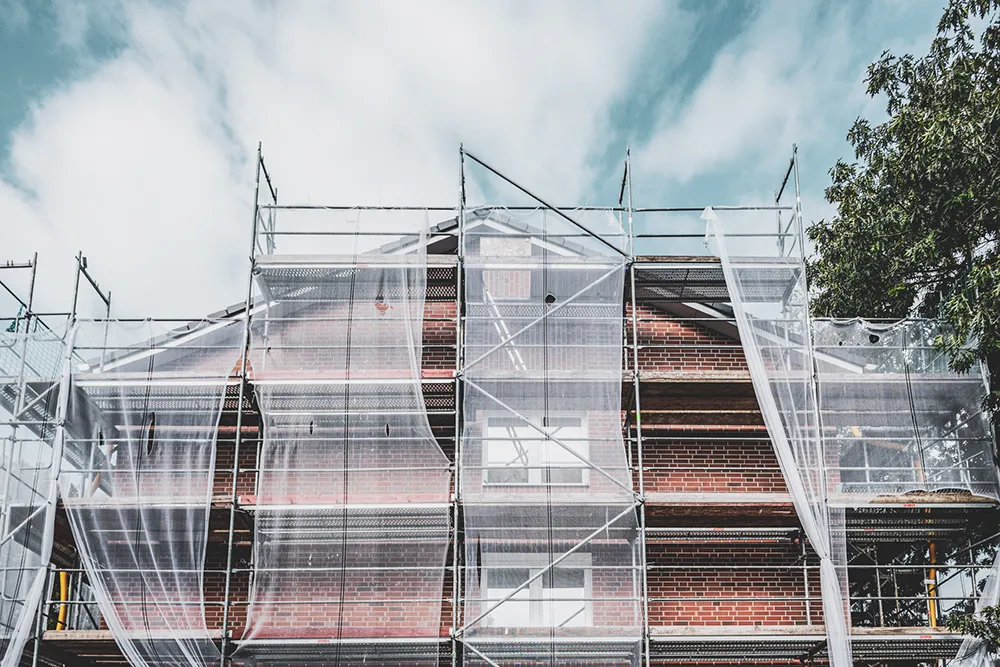 Malerei Juergens aus Hamburg sorgt mit einem sicheren und geprüften Baugerüst vor der roten Steinziegelwand für reibungsloses Arbeiten, den Schutz unserer Mitarbeiter sowie Ihres Eigentums. Weiße Netze hängen von der obersten Ebene bis nach unten. Das Baugerüst steht nicht nur für Sicherheit, sondern auch symbolisch für den Schutz Ihrer Daten.