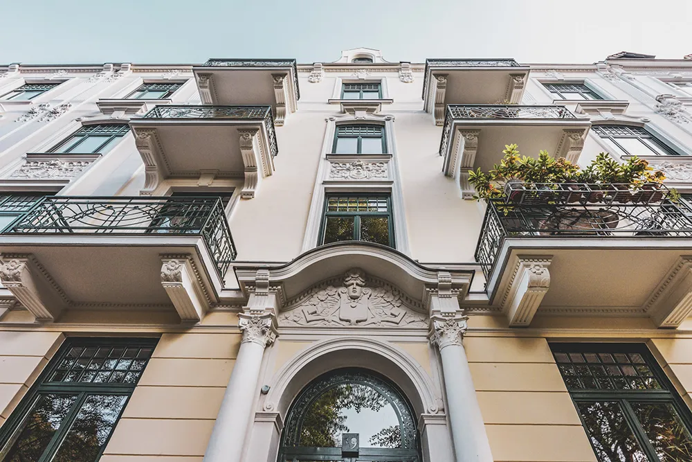 Ein Blick auf ein Hamburger Wohnhaus, das von Maler Juergens mit Schmucktechniken für Balkone und Fassade gestaltet wurde. Der Blick fällt von unten auf das Gebäude, wobei links und rechts des Bildzentrums an jedem Stockwerk einheitliche Balkone zu sehen sind. Die Fassade und ihre Schmuckelemente erstrahlen in einem cremefarbenen Ton, während die Balkongitter und Fenster in einem eleganten Dunkelgrün hervorstechen.