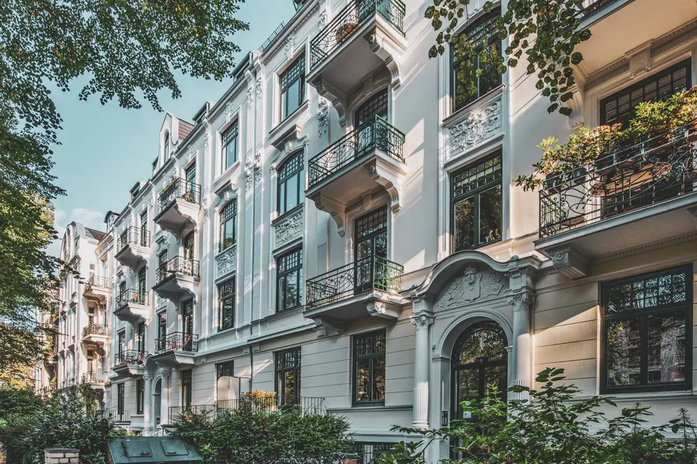 Auf dem Bild ist ein Referenzobjekt in Hamburg von Maler Juergens zu sehen. Eine ästhetisch ansprechende cremefarbene Hausfassade mit hochwertigen Schmuckelementen. Die Fensterrahmen, Türrahmen und Balkone sind in einem dunklen Grün gestaltet. Grüne und blühende Pflanzen sorgen für ein natürliche Balance aus Handwerkskunst und Natur.