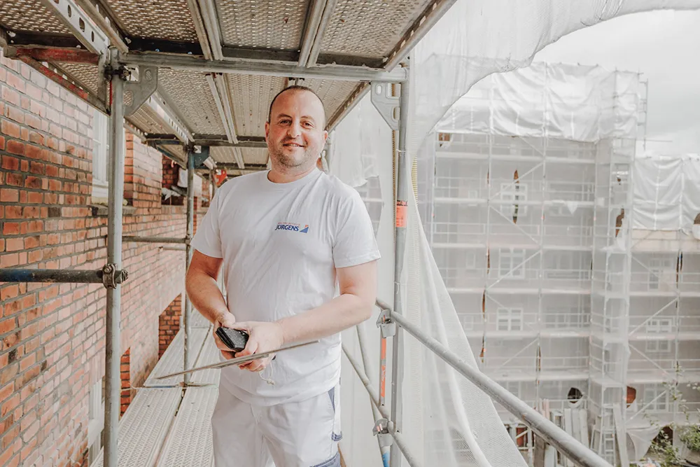 Ein Mitarbeiter von Malerei Juergens steht auf einem Baugerüst vor einer roten Ziegelsteinwand in Hamburg und lächelt in die Kamera. Er trägt ein weißes Arbeitsshirt und eine weiße Arbeitshose und hält einen Spatel in den Händen. Das Bild symbolisiert eine freundliche Kontaktaufnahme mit Malerei Juergens – wir stehen bereit, Ihre Wünsche professionell umzusetzen.